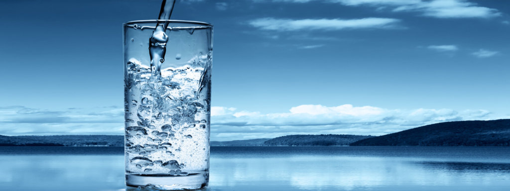 Water being poured into a clear glass in front of a beautiful blue lake and sky.